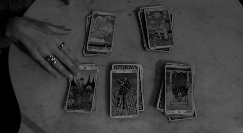 Black-and-white overhead shot of a tarot card reading: five tarot piles on a marble table with a ringed hand touching one card