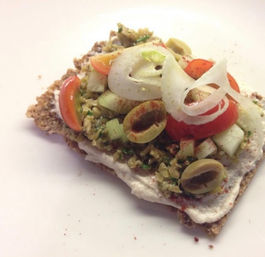 Crunchy seed crispbread topped with creamy spread, chopped olive tapenade, cucumber cubes, cherry tomato halves, sliced green olives, thin onion rings and a dusting of paprika on a white plate — Mediterranean-style snack.