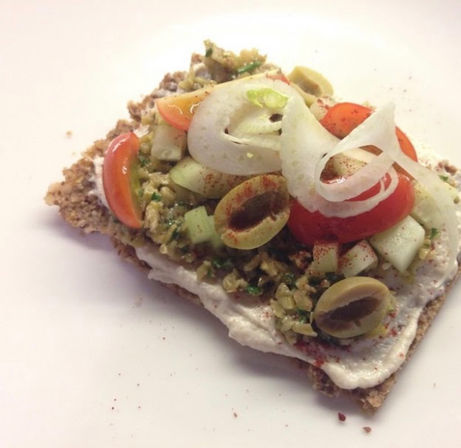 Crunchy seed crispbread topped with creamy spread, chopped olive tapenade, cucumber cubes, cherry tomato halves, sliced green olives, thin onion rings and a dusting of paprika on a white plate — Mediterranean-style snack.