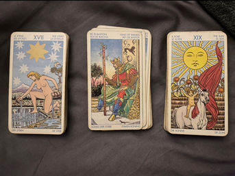 Flat lay of three illustrated tarot card stacks on dark fabric: The Star (XVII) showing a pouring figure, King of Wands seated with staff, and The Sun (XIX) with a child on a white horse.