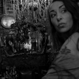 Moody black-and-white portrait of a woman glancing sideways inside a dim vintage shop with ornate mirrors, a crystal chandelier and dried roses.