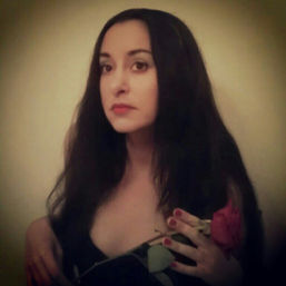 Moody indoor close-up portrait of a woman with long dark hair and red lipstick holding a red rose near her chest against a neutral background, soft vignette and warm lighting.