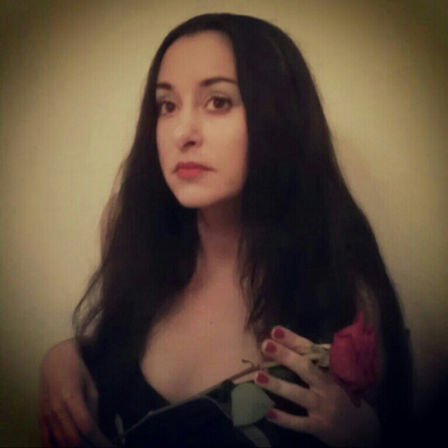 Moody indoor close-up portrait of a woman with long dark hair and red lipstick holding a red rose near her chest against a neutral background, soft vignette and warm lighting.