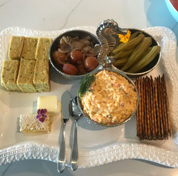 Appetizer platter on a white decorative tray: creamy pimento cheese dip with dill in a seahorse three-compartment bowl, pickled cherry tomatoes, pickled okra, cornbread squares, honeycomb with butter and edible flower, stacked crackers and serving spoons.