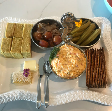 Appetizer platter on a white decorative tray: creamy pimento cheese dip with dill in a seahorse three-compartment bowl, pickled cherry tomatoes, pickled okra, cornbread squares, honeycomb with butter and edible flower, stacked crackers and serving spoons.
