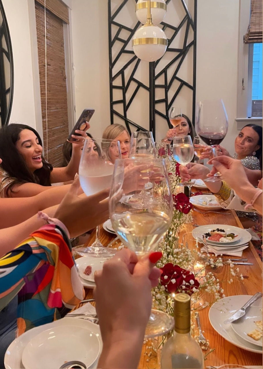 Friends toasting with wine glasses at a cozy indoor dinner party around a long wooden table decorated with roses and baby's breath.