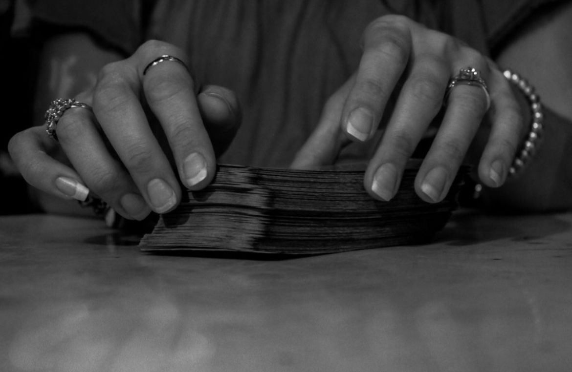 Close-up black-and-white image of hands with rings and a beaded bracelet handling a worn deck of cards on a tabletop