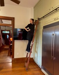 Person posing on a rolling library ladder to access high pale-green kitchen cabinets beside a stainless-steel refrigerator, hardwood floors and wall-mounted TV.