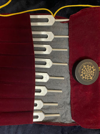 Eight metal tuning forks neatly arranged in a red velvet roll-up pouch with a yellow cord, accompanied by a black tin with a gold geometric design — sound therapy and meditation tools on dark fabric.