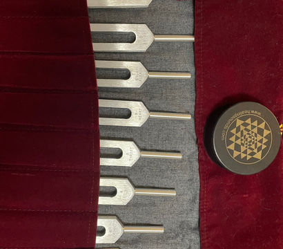 Eight metal tuning forks neatly arranged in a red velvet roll-up pouch with a yellow cord, accompanied by a black tin with a gold geometric design — sound therapy and meditation tools on dark fabric.