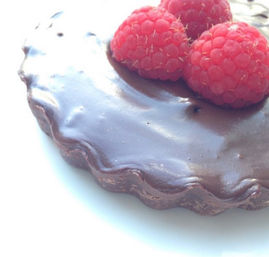 Close-up of a glossy chocolate ganache tartlet topped with three fresh raspberries on a white plate
