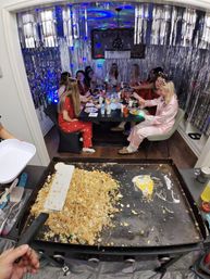Indoor pajama party in a silver-foil, disco-lit dining nook with people in colorful pajamas at a table while a cook flips fried rice on a large griddle in the foreground.