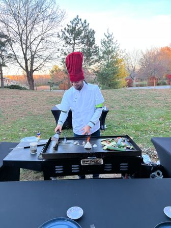 Chef in a tall red toque and white jacket cooking vegetables on a flat-top griddle outdoors in an autumn backyard, spatula in hand with a small flame on the grill and plates on a table in the foreground.