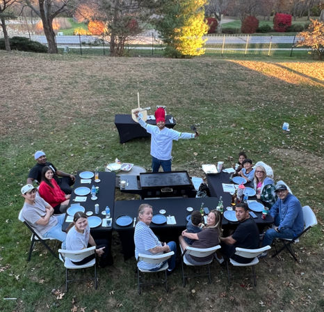 Backyard autumn family barbecue with a chef wearing a red chef hat and holding a spatula at a central griddle, surrounded by a multi-generation group seated at U-shaped black tables on the lawn with trees and a road in the background.