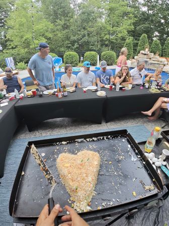 Poolside backyard party with guests seated at a long table while hands on a griddle shape a large heart-shaped fried rice in the foreground — outdoor summer cookout.