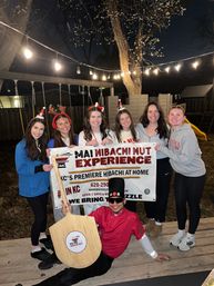 Outdoor backyard party at night: six smiling friends wearing holiday headbands pose with a hibachi chef holding a large wooden paddle in front of a promotional banner under string lights on a wooden deck.