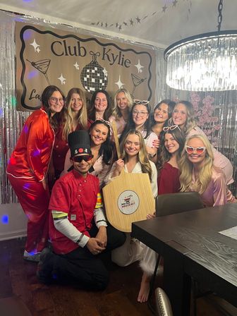 Dozen women in pink and red pajamas smiling in a decorated dining room for a girls’ night/bachelorette-style party with silver fringe backdrop, disco-ball banner, chandelier, and a chef in red holding a wooden peel.
