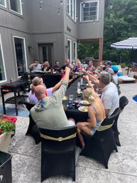 Cheerful backyard patio party at a modern gray house: a large group around a long black-covered table raising cups for a toast under string lights, with poolside umbrella and trees in the background.