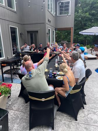 Cheerful backyard patio party at a modern gray house: a large group around a long black-covered table raising cups for a toast under string lights, with poolside umbrella and trees in the background.