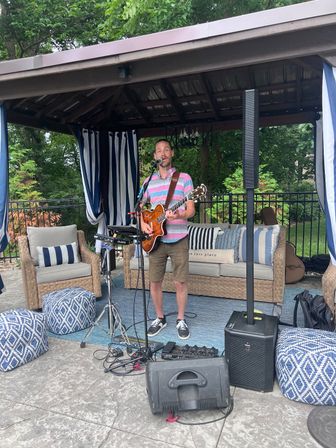 Solo musician strumming a hollow-body guitar and singing under a backyard gazebo, surrounded by wicker patio seating, blue-and-white striped cushions, patterned poufs and portable speakers.