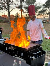 Outdoor hibachi-style chef wearing a tall red toque and white jacket cooking on a flat-top grill as towering orange flames leap up in a backyard at sunset.