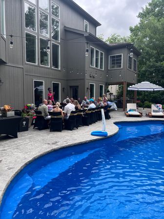 Backyard poolside party at a modern two-story gray house — long outdoor dining table with guests under string lights on a stamped concrete patio, bright blue in-ground pool, striped umbrella and lounge chairs.