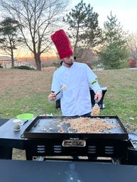 Chef in white coat with tall red hat flipping fried rice on a large flat-top griddle outdoors in a backyard at sunset, trees and houses in the background.