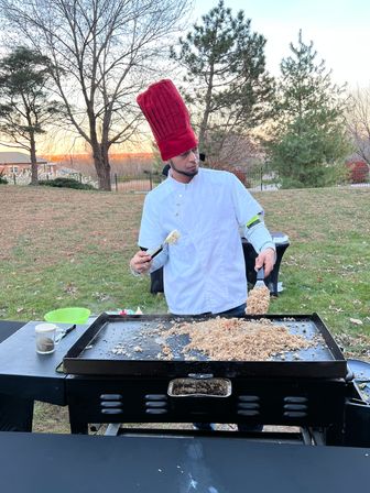 Chef in white coat with tall red hat flipping fried rice on a large flat-top griddle outdoors in a backyard at sunset, trees and houses in the background.