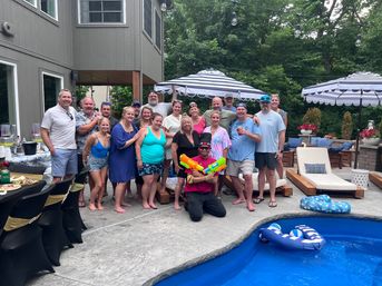 Backyard summer pool party group photo on a patio with striped umbrellas, lounge chairs and blue pool floats; a person kneeling front holds colorful water guns while friends pose around the pool.