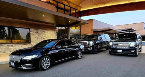 Black luxury sedan and two black SUVs parked under a modern hotel's wood-panecked porte-cochere at dusk, warm exterior lighting and stone facade suggesting a valet-ready entrance.