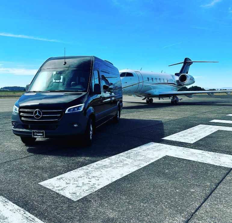 Black luxury shuttle van parked on an airport tarmac beside a white private jet, runway markings in the foreground and clear blue sky overhead.
