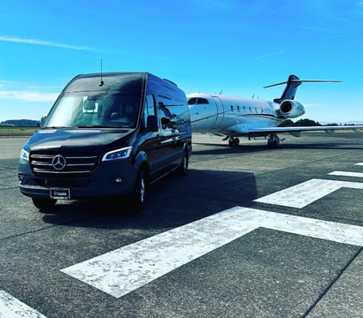 Black luxury shuttle van parked on an airport tarmac beside a white private jet, runway markings in the foreground and clear blue sky overhead.