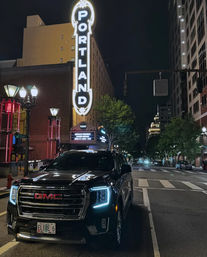 Night in downtown Portland, Oregon — black GMC SUV with glowing LED headlights parked beneath the iconic vertical "PORTLAND" marquee, lit buildings and empty street.