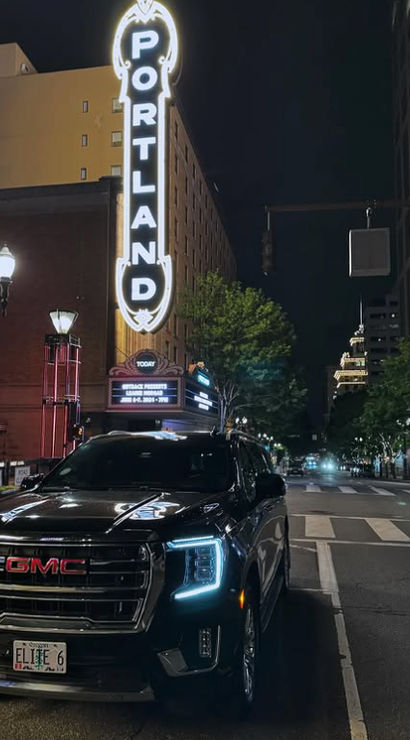 Night in downtown Portland, Oregon — black GMC SUV with glowing LED headlights parked beneath the iconic vertical "PORTLAND" marquee, lit buildings and empty street.