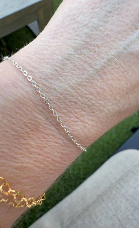 Close-up of a wrist wearing a delicate silver chain bracelet and a chunky gold chain bracelet, photographed outdoors on a patio with green grass in the background.
