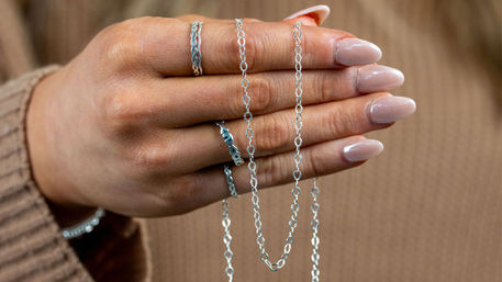 Close-up of a hand with glossy nude manicure holding delicate linked silver chain necklaces, wearing stackable silver rings against a beige knit sweater backdrop