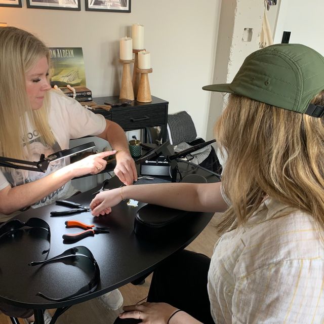 Hands-on home jewelry studio scene — a technician adjusts a bracelet on a seated woman’s wrist at a black work table with pliers, safety glasses, magnifying lamp and decorative candles in the background.