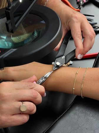 Close-up of hands using needle-nose pliers under a magnifying lamp to precisely repair a delicate gold chain bracelet on a wrist at a jewelry workbench