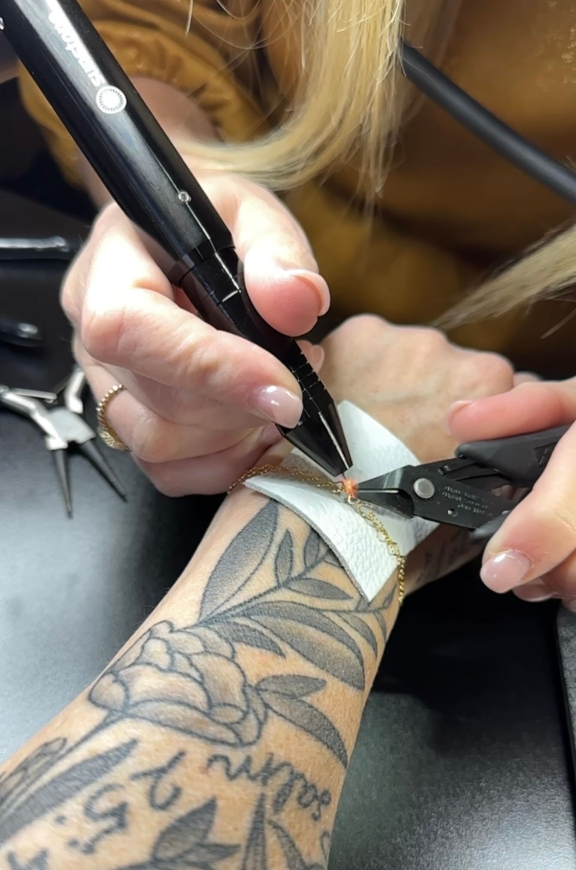 Close-up of hands repairing a delicate gold chain bracelet on a tattooed forearm, using needle-nose pliers and a black handheld jewelry tool over a white pad.