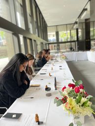 Participants at a hands-on workshop seated along a long white-covered table in a bright glass-walled modern event space, with worksheets, notebooks, pens and small amber bottles on the table and a red-and-white floral centerpiece in the foreground.