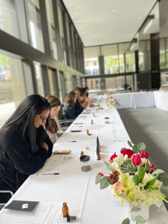 Participants at a hands-on workshop seated along a long white-covered table in a bright glass-walled modern event space, with worksheets, notebooks, pens and small amber bottles on the table and a red-and-white floral centerpiece in the foreground.