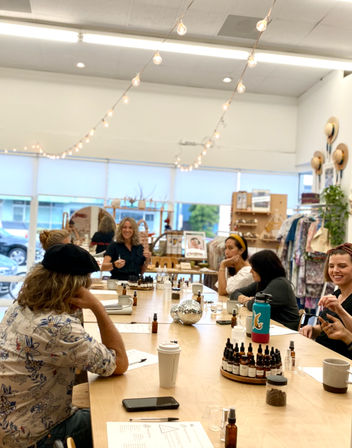 Group of adults at an essential-oil blending workshop in a bright boutique studio — seated around a long wooden table with amber dropper bottles, sample vials, coffee cups, string lights overhead and clothing racks along the wall.