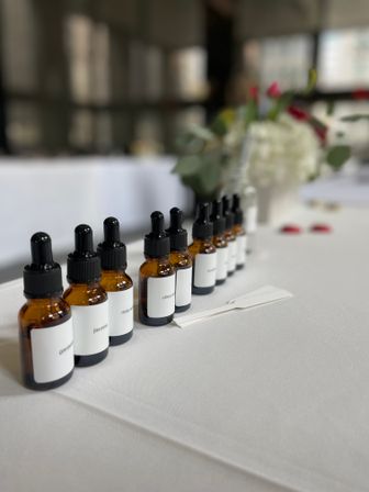 Amber glass dropper bottles labeled jasmine, rose and citrus lined up on a white tablecloth for an indoor fragrance tasting, with a blurred floral centerpiece in the background.
