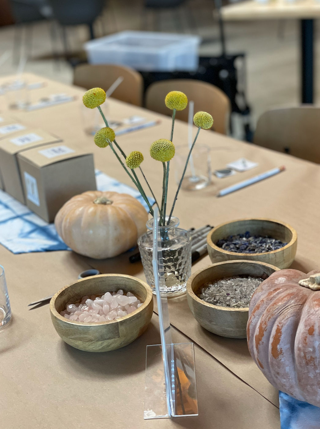 Indoor craft workshop table with yellow billy ball flowers in a glass vase, pumpkins and wooden bowls filled with rose quartz, clear crystals and decorative stones.