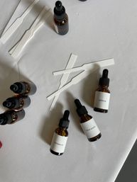 Flat-lay of amber glass dropper bottles labeled sandalwood, citrus accord and gardenia with paper scent strips on a white tablecloth — fragrance sampling setup.