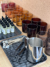 Candle-making setup on a wooden table with rows of amber and dark brown glass candle jars, a stainless-steel pouring pitcher, pre-tabbed cotton wicks, black wick holders, and small dropper bottles of fragrance oil arranged on trays.