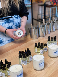 Participant at a DIY candle-making workshop holding a floral-topped soy candle above a wooden workbench lined with fragrance oil droppers, stainless steel pouring pitchers, and finished glass candles in an artisan studio