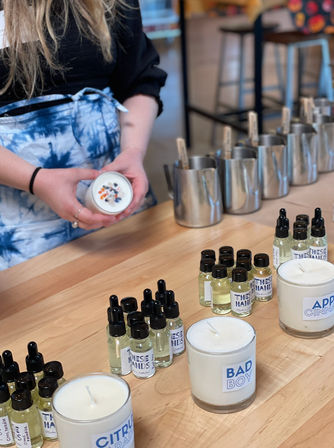 Participant at a DIY candle-making workshop holding a floral-topped soy candle above a wooden workbench lined with fragrance oil droppers, stainless steel pouring pitchers, and finished glass candles in an artisan studio