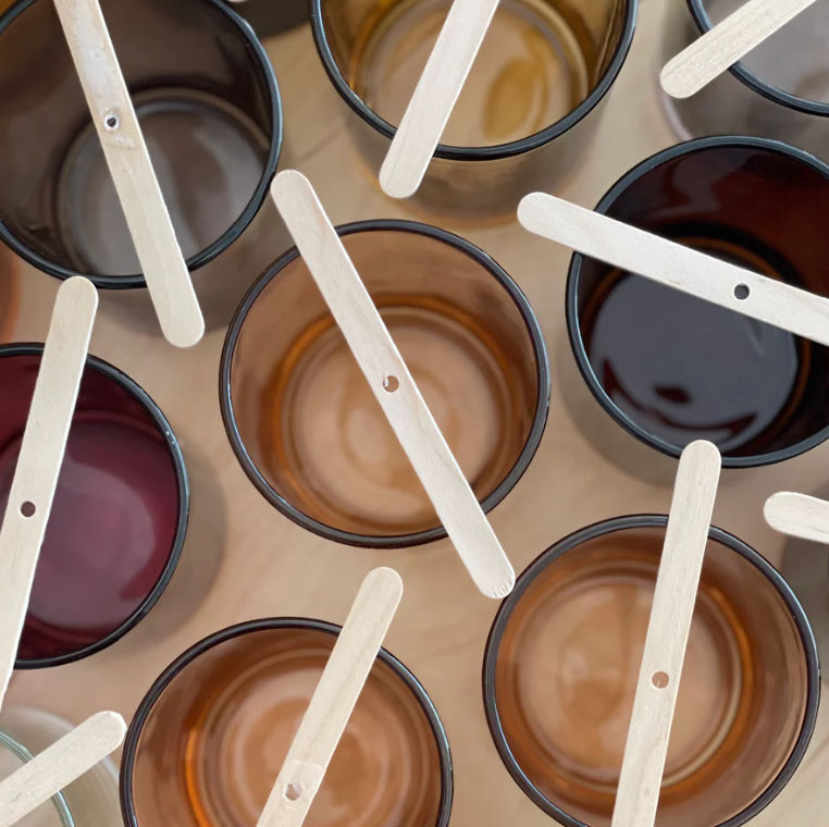 Top-down flat lay of amber and dark glass jars with wooden craft sticks laid across them, a cozy candle-making scene.