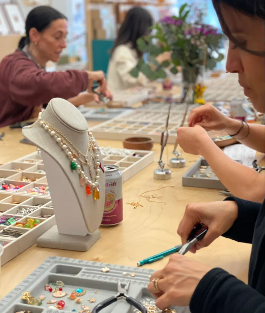 Close-up of a handmade jewelry workshop: crafters' hands using pliers at a wooden table with bead trays, tools, and a display bust showing layered necklaces.